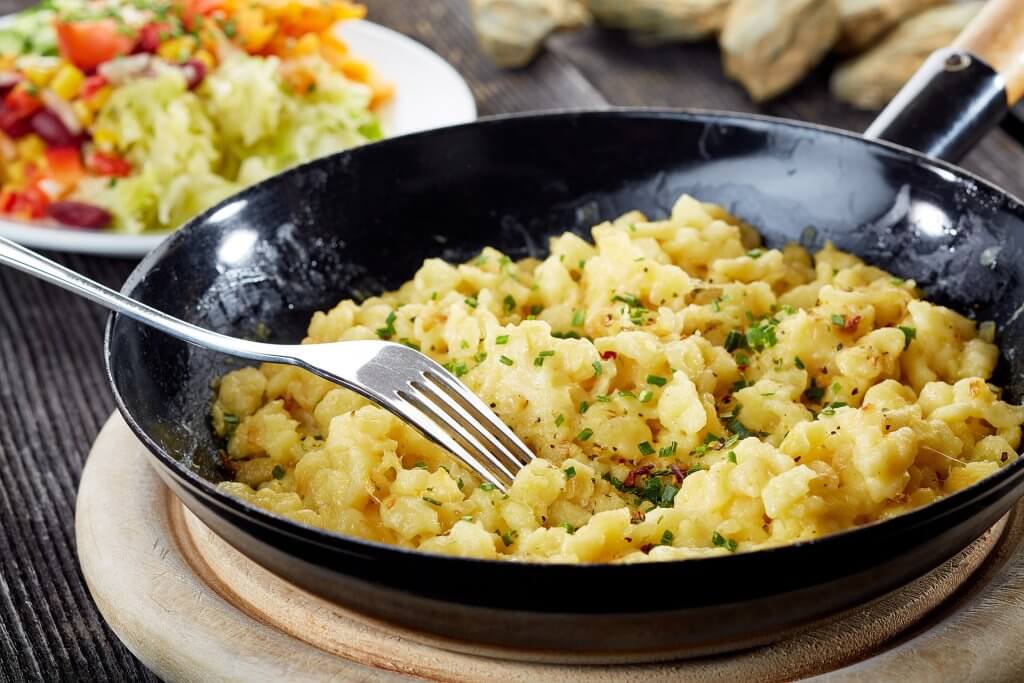 Kasnocken aus WOERLE Käse stehen auf einem Holzbrett, in einer Pfanne auf dem Tisch. Im Hintergrund sieht man einen Salat.