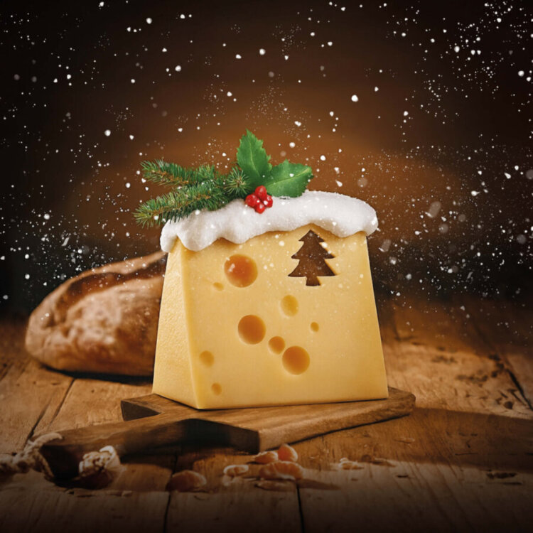 Emmentaler mit einem Loch in Form eines Baumes. Auf dem Käse schneit es, sodass sich eine Schneedecke auf ihm gebildet hat.