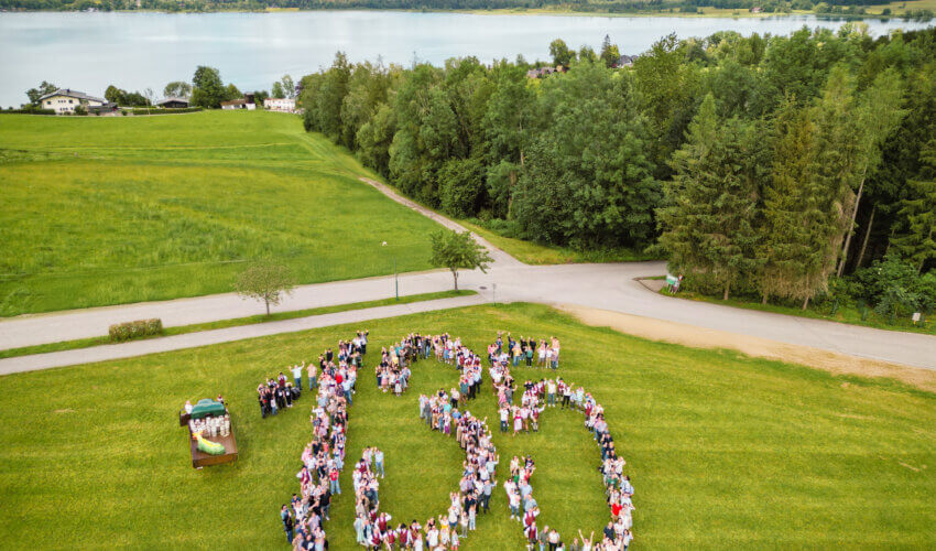 WOERLE-Mitarbeitende formen die Zahl 135 auf einer Wiese zum 135-jährigen Jubiläum – im Hintergrund der Wallersee in Henndorf