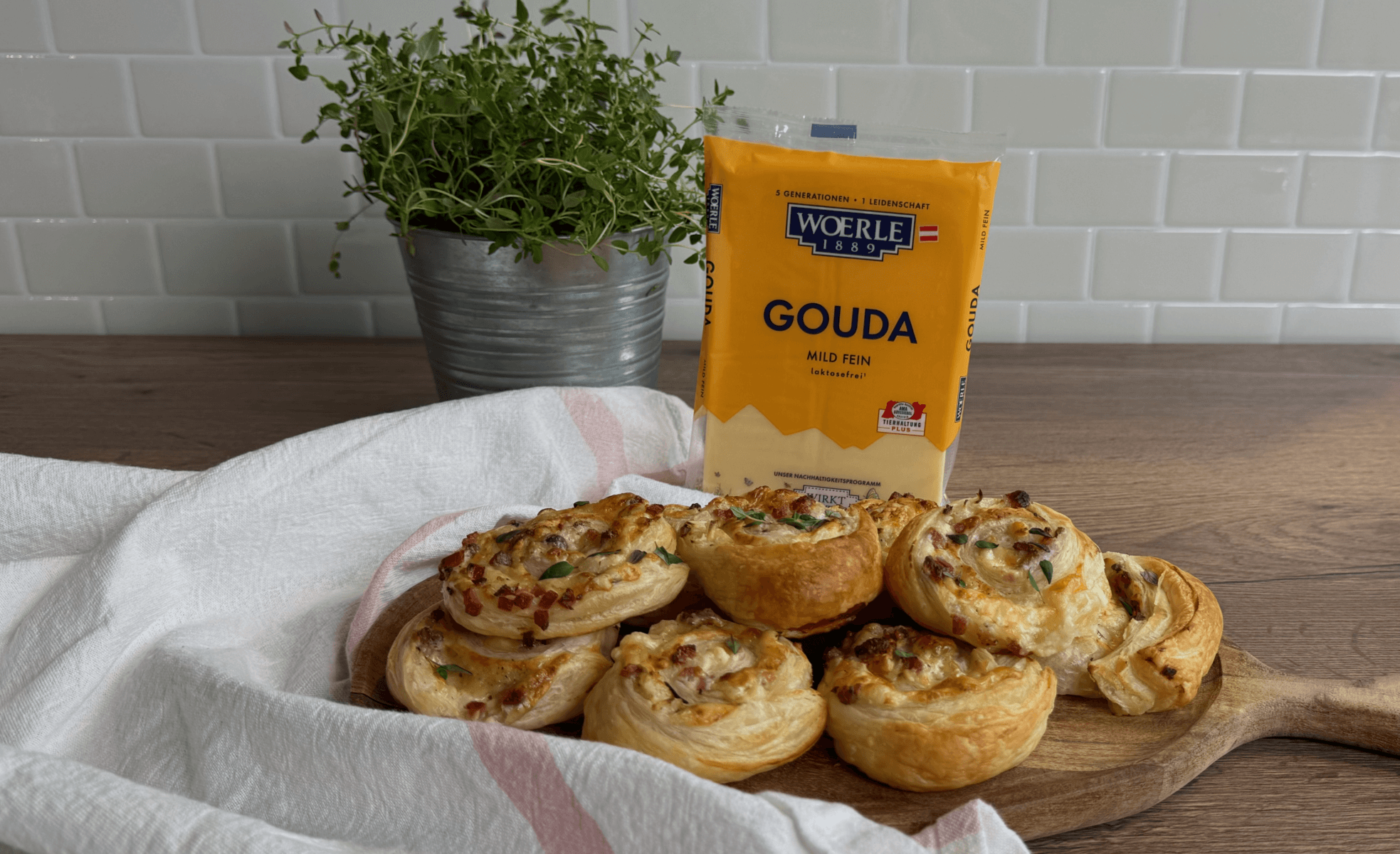 Ein Rezept für herzhafte Flammkuchen-Schnecken mit WOERLE Gouda. Die goldbraun gebackenen Blätterteigschnecken sind gefüllt mit würzigem Käse, Speck und Frühlingszwiebeln. Serviert werden sie auf einem rustikalen Holzbrett, das auf einer weißen Stoffserviette liegt.
