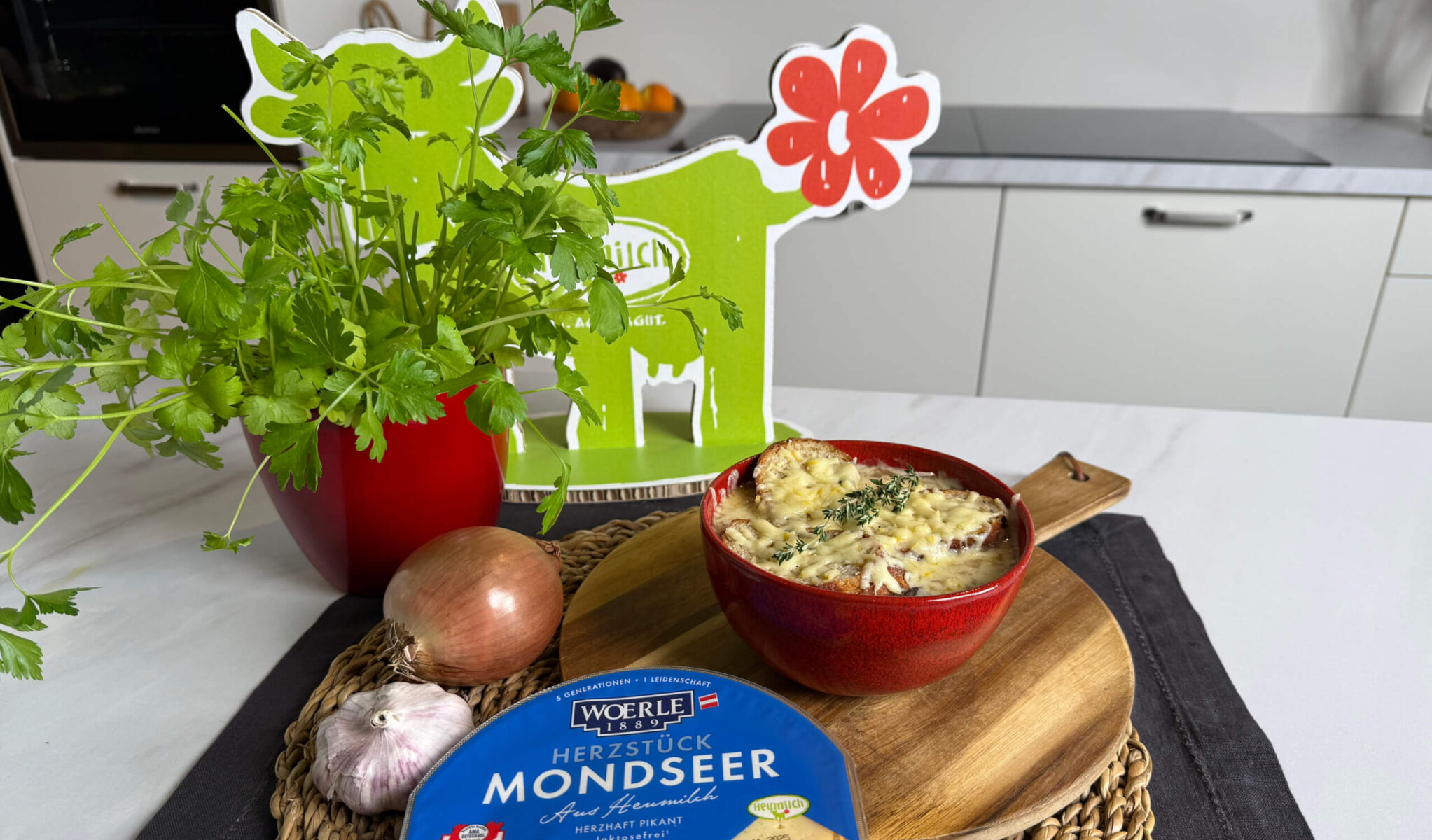 Auf einer hellen Küchenarbeitsfläche steht eine Schale Zwiebelsuppe mit gratiniertem Käse und frischem Thymian, daneben Zwiebel, Knoblauch, Petersilie im roten Topf und eine Packung WOERLE Mondseer aus Heumilch, arrangiert vor einer grünen Kuh-Dekoration.